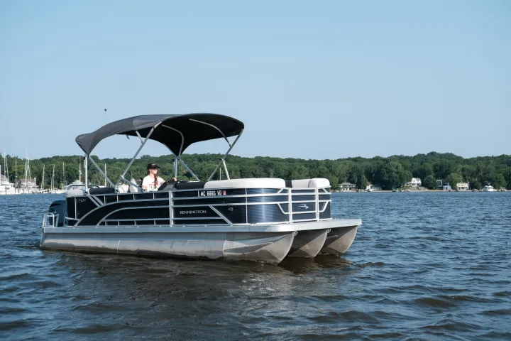 Man on a pontoon boat with canopy on a lake, distant shore with trees.