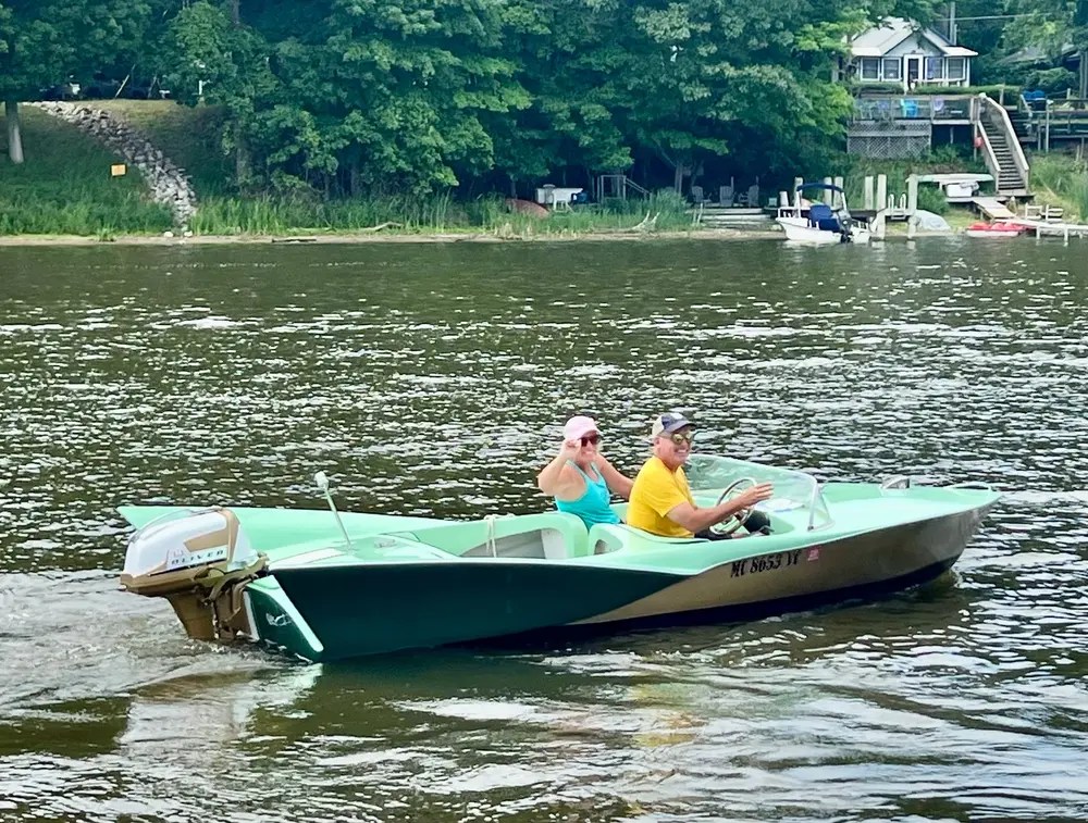 Two people on a small boat in a river near a tree-lined shore.
