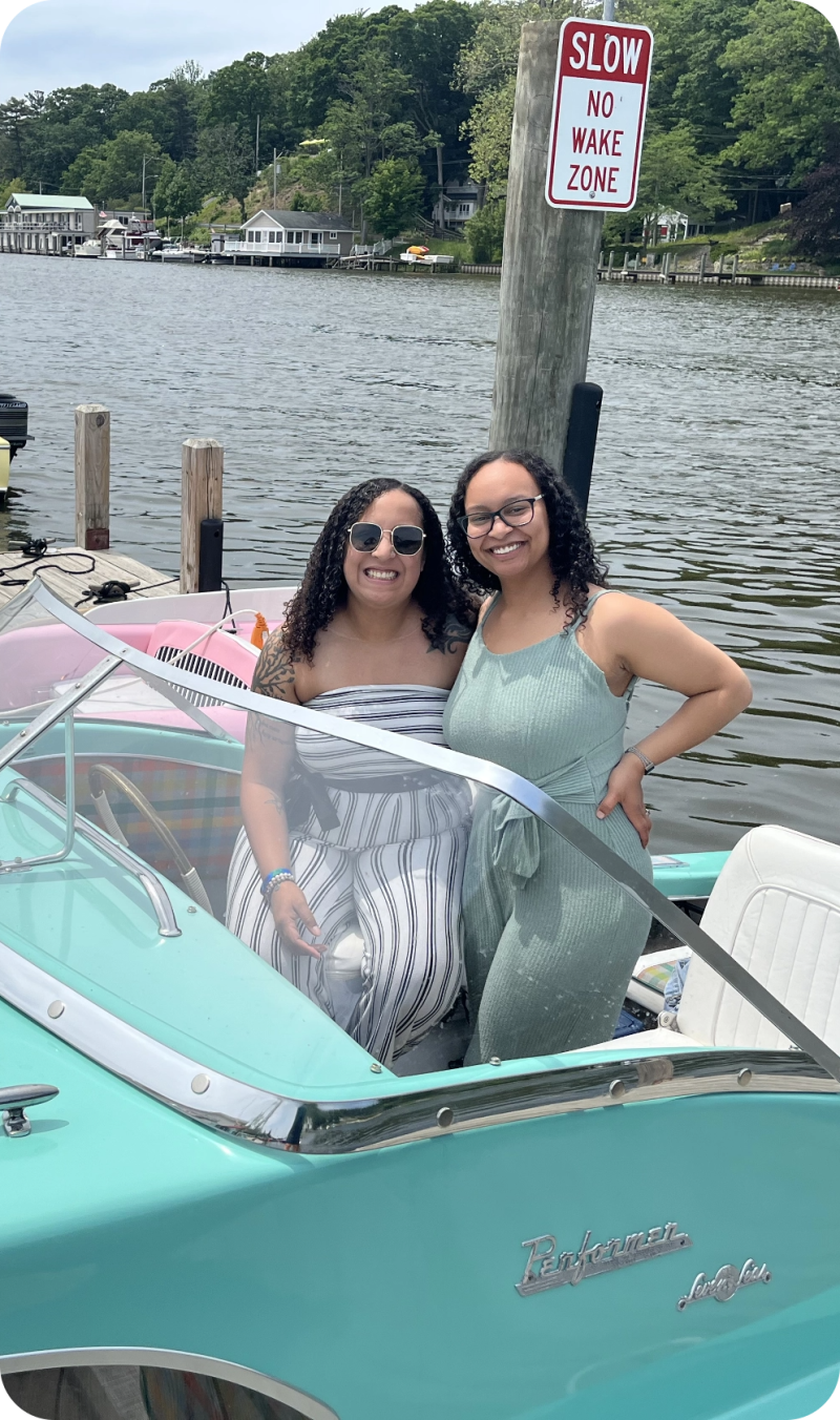 Two smiling women in a boat by a lake with a 'No Wake Zone' sign.