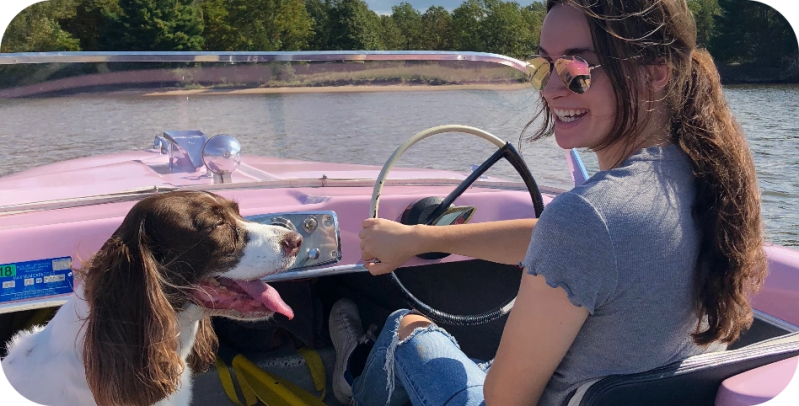 Woman driving pink boat with a dog on a lake, both smiling.