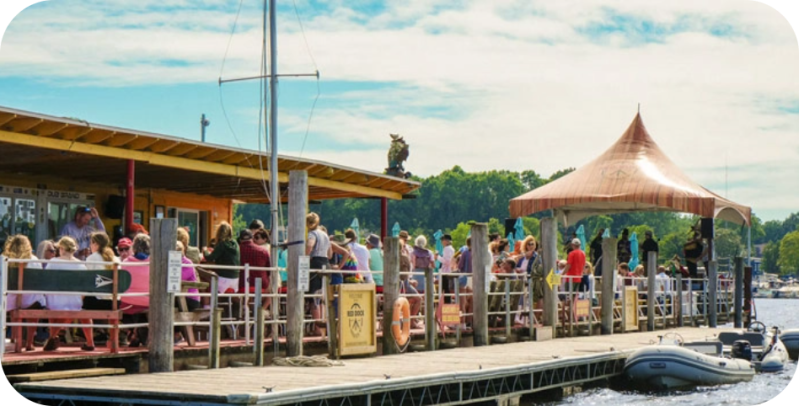 Crowded dock with people sitting and standing, under a tent on a sunny day.