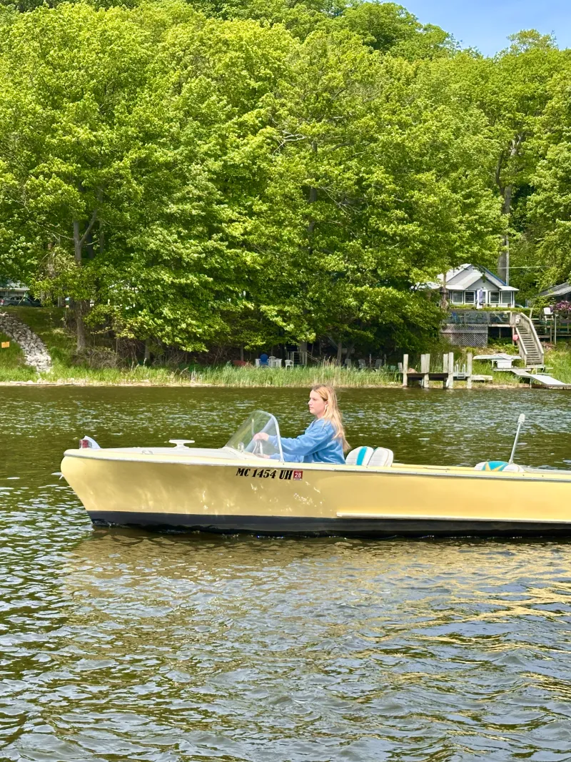 Yellow motorboat on a lake with a person steering; green trees and houses in the background.